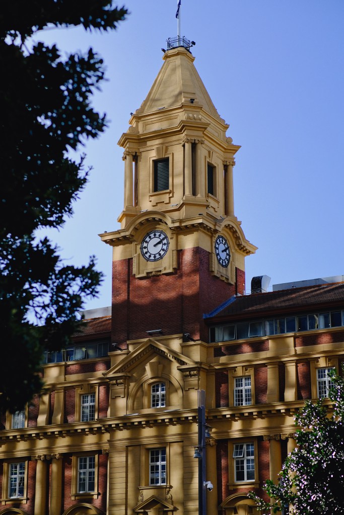 Historic building featuring a tall clock tower with a peaked roof and decorative details, set against a blue sky.