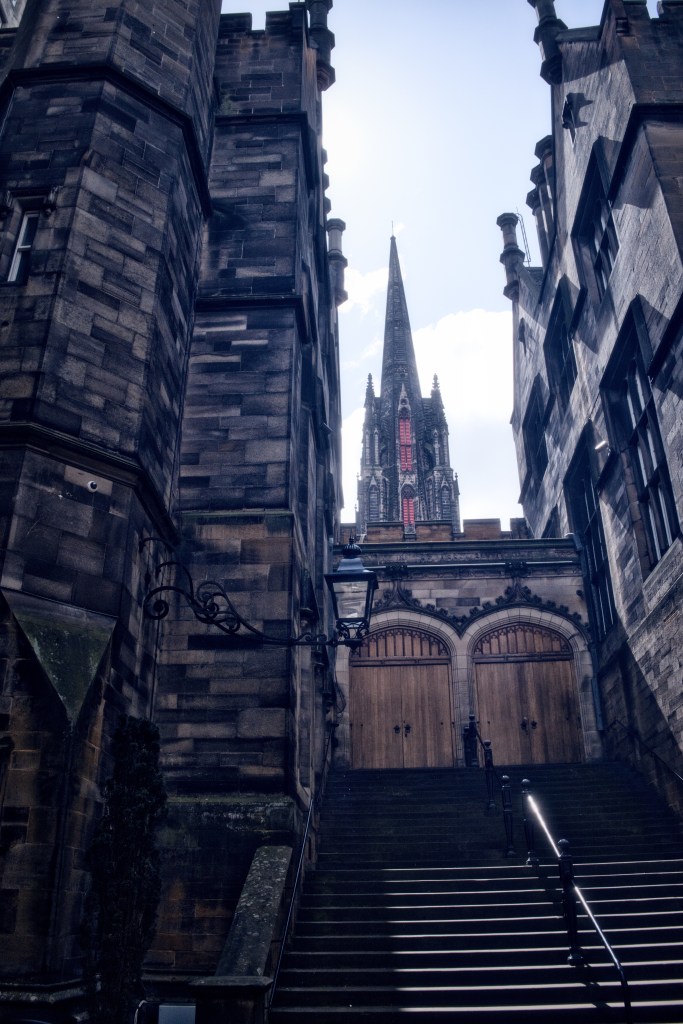 View of a narrow alleyway between historic stone buildings, leading to a wooden door and a tall church spire in the background under a cloudy sky.