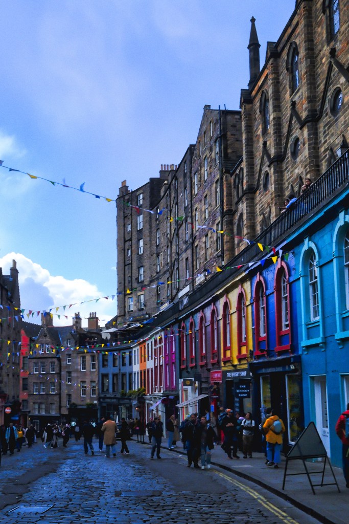 A vibrant street in Edinburgh featuring colorful buildings, cobblestone pavement, and people walking. Decorative bunting hangs overhead against a blue sky.