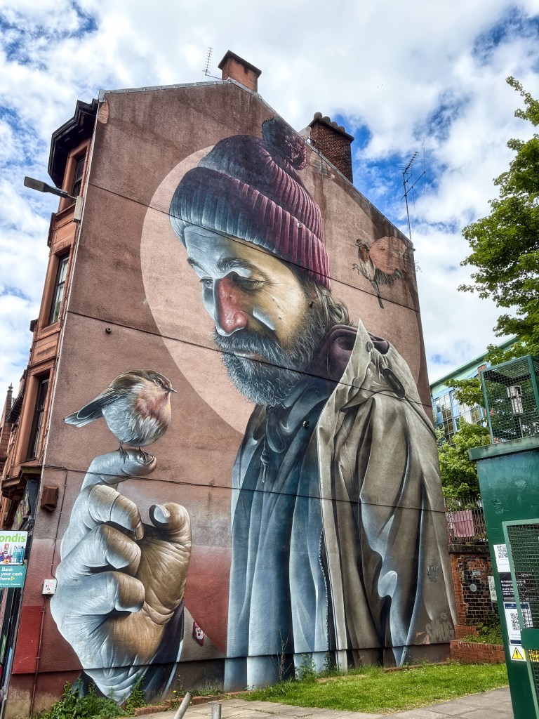 A large mural depicting an elderly man with a beard wearing a beanie, holding a small bird on his finger, against a cloudy sky backdrop.