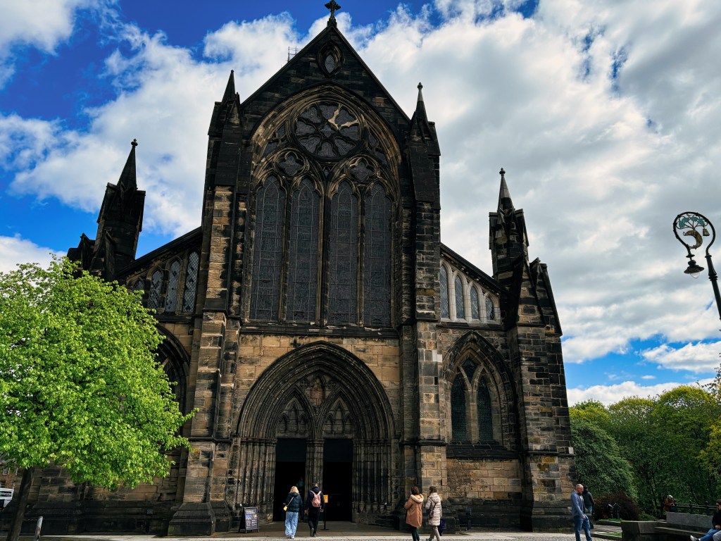 Historic church building with Gothic architecture under a partly cloudy sky, surrounded by greenery and people walking.
