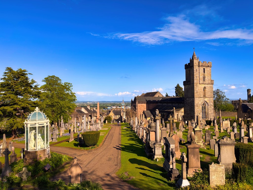 A panoramic view of a cemetery with ancient gravestones, lush greenery, and a historic church tower under a clear blue sky.