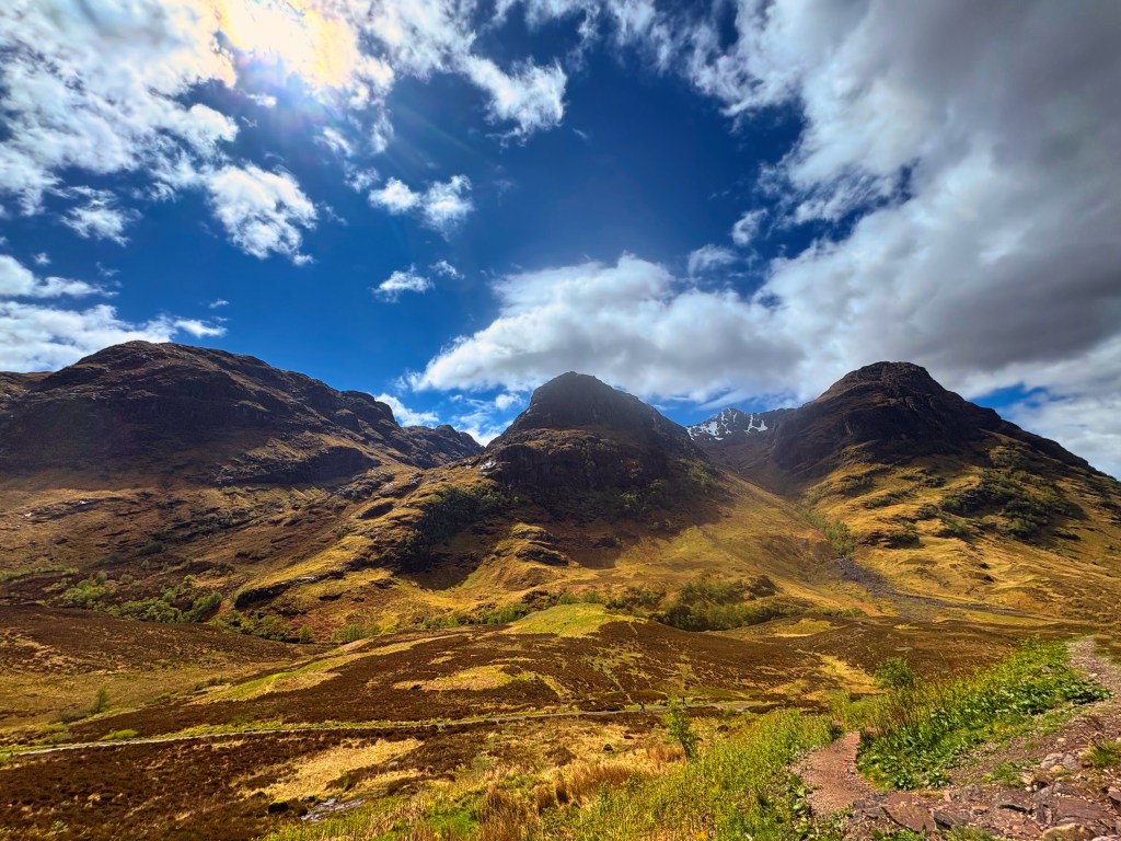 A scenic view of rugged mountains under a partly cloudy sky, with patches of green and brown landscape in the foreground.