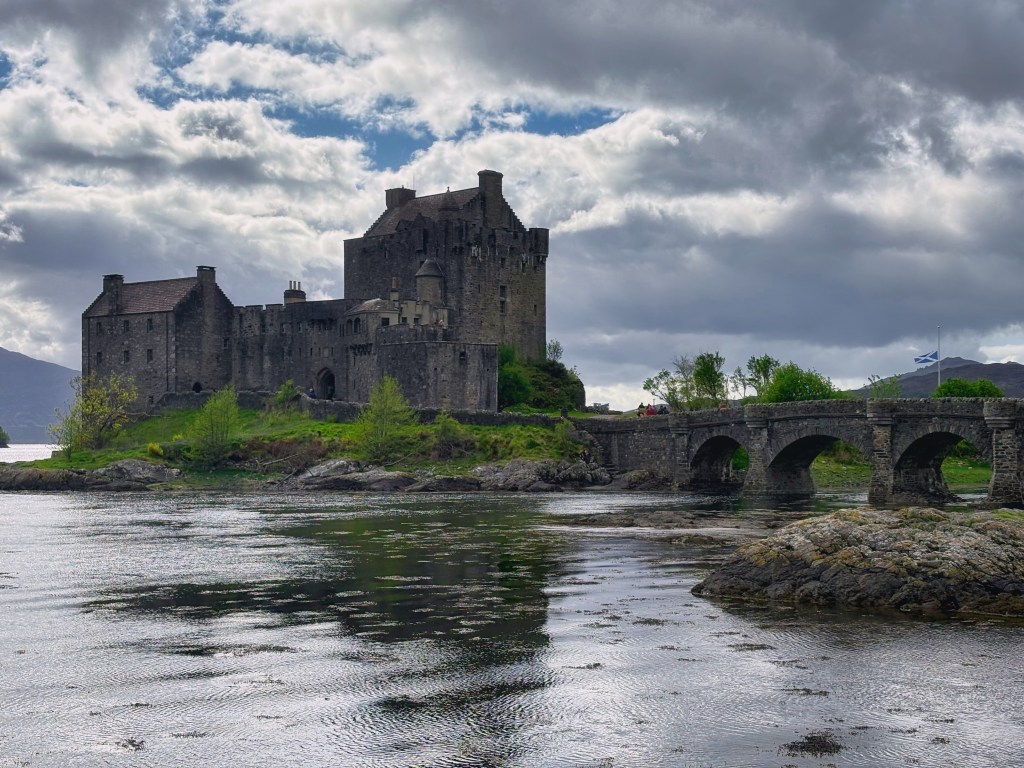 Historic castle situated on an island, surrounded by water and greenery, with a stone bridge leading to it. The sky is cloudy, enhancing the dramatic scenery.