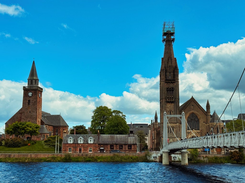 A scenic view of historic buildings with clock towers, set alongside a river and a bridge, under a partly cloudy sky.