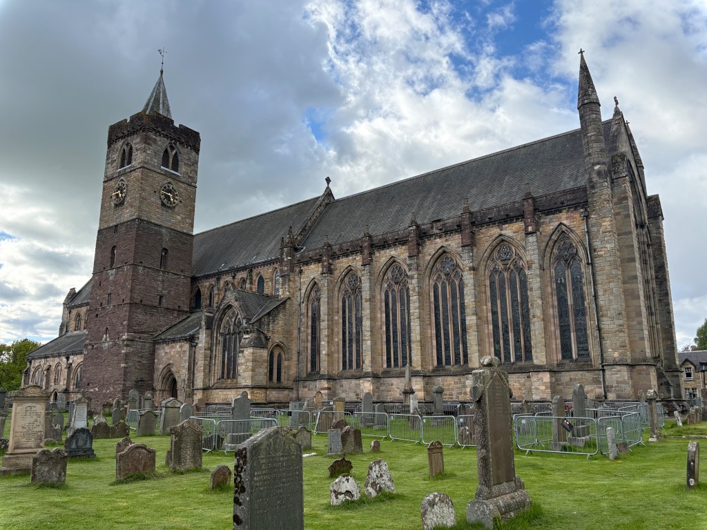 A large stone church featuring a tall bell tower, surrounded by a graveyard with various headstones and grassy areas, under a partly cloudy sky.