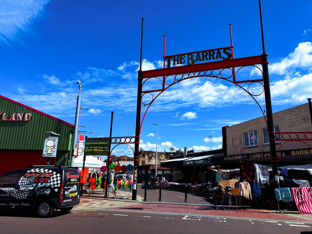 Entrance arch of The Barras market with vibrant blue sky, various stalls, and vehicles parked in front.