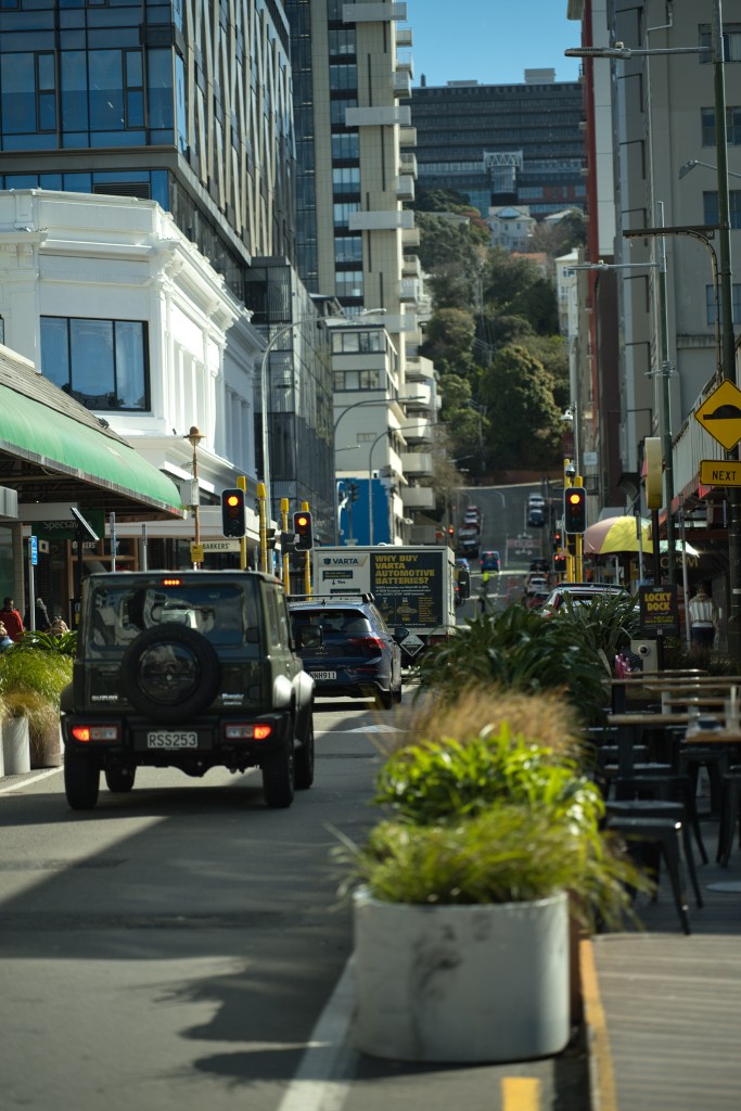 A city street scene featuring a green SUV driving past outdoor seating areas with potted plants and various storefronts under a clear blue sky.