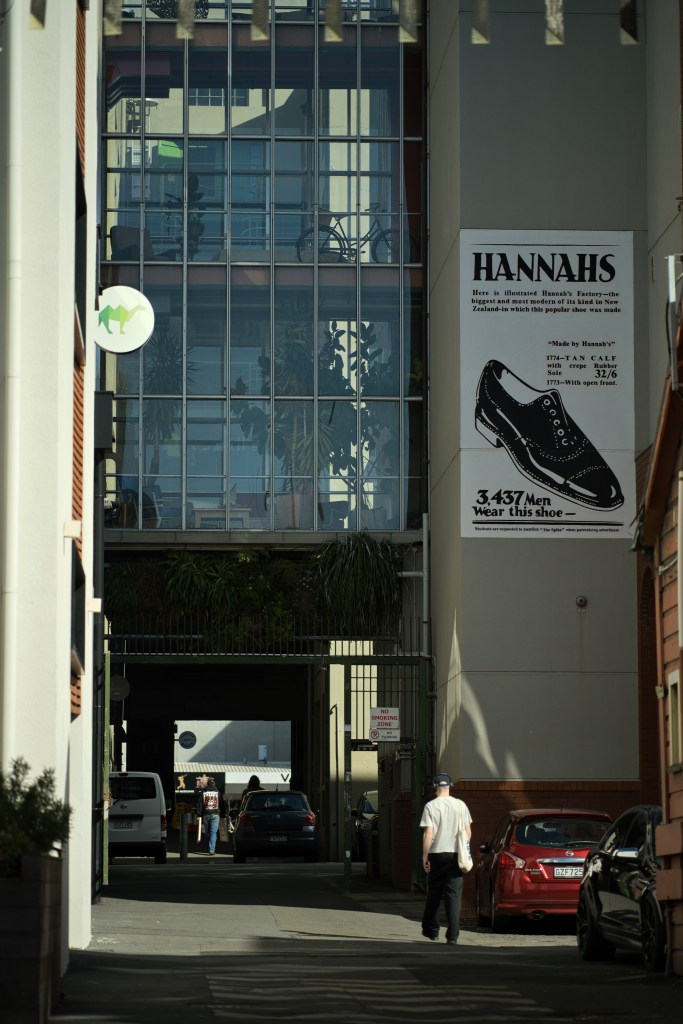 View of a street with a person walking towards an entrance, featuring a large advertisement for Hannah's shoe store on the wall.