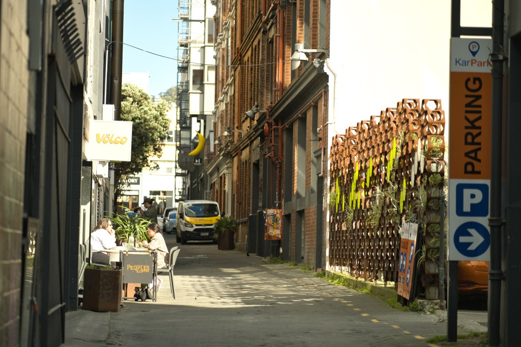 A narrow alleyway lined with brick buildings, featuring a café table with two patrons and greenery. A parking sign is visible on the right, and a van is parked nearby.