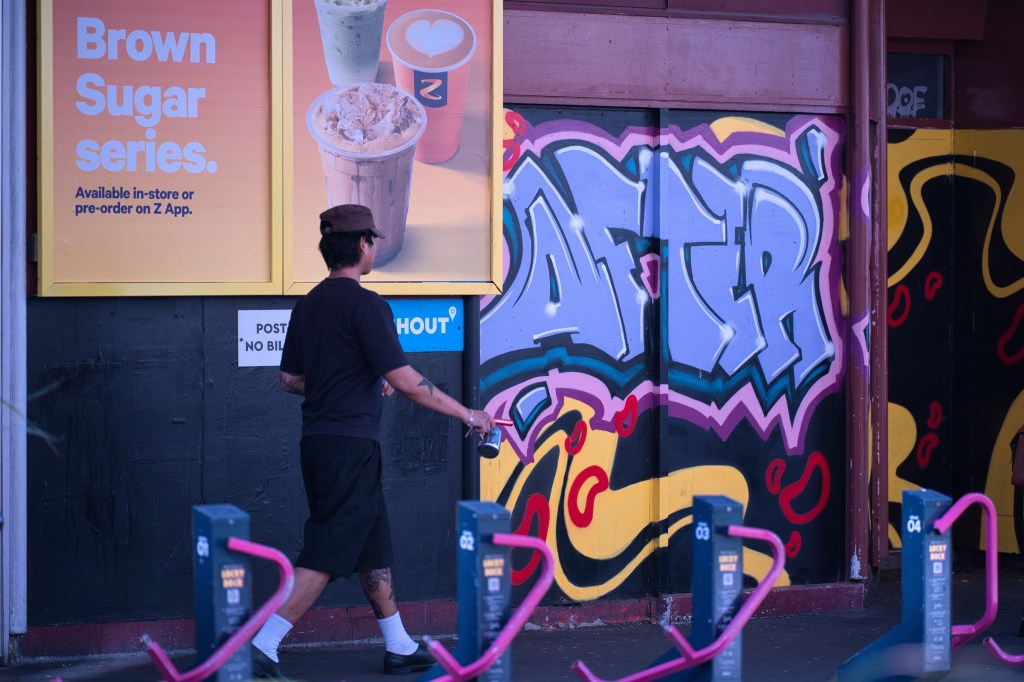 A person walking past a colorful graffiti wall featuring the word 'AFTER'. There is a promotional poster for a drink series titled 'Brown Sugar' on the left, showcasing various drink options. The scene includes bike racks in the foreground with bright pink designs.