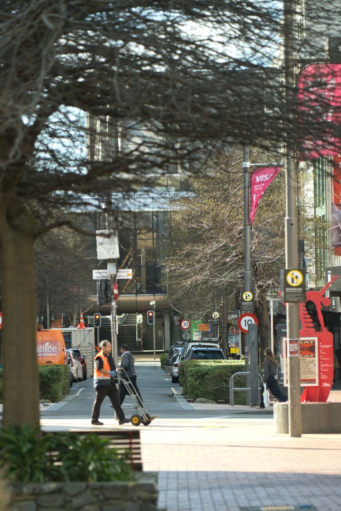 A city street scene featuring a man in an orange vest pushing a hand truck, with traffic lights and parked cars lining the road. In the background, there are modern buildings and street signs visible.