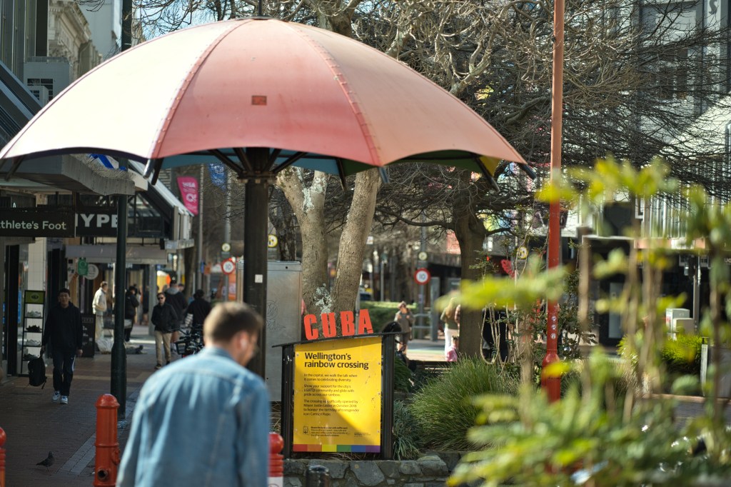 Colorful umbrella in a street area with shops, featuring a sign about Wellington's rainbow crossing.