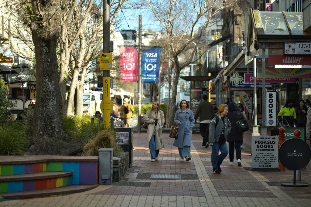 A busy street scene featuring pedestrians walking on a brick pavement, surrounded by shops and cafes, with trees lining the walkway and colorful seating along the side.