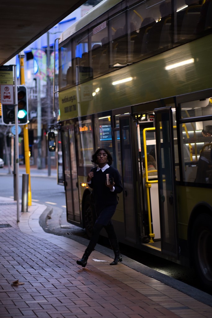 A person in dark clothing hurries past a yellow bus at a bus stop in a city setting during twilight.