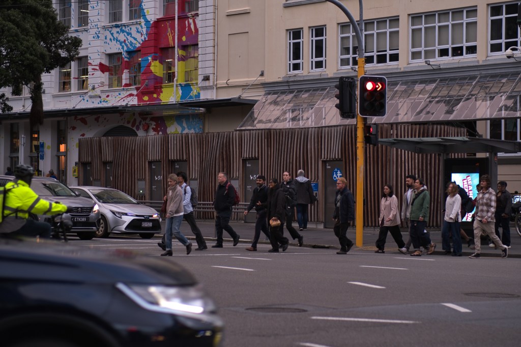 A group of pedestrians crossing the street at a traffic light, with vehicles in the foreground and a colorful mural on the building behind them.