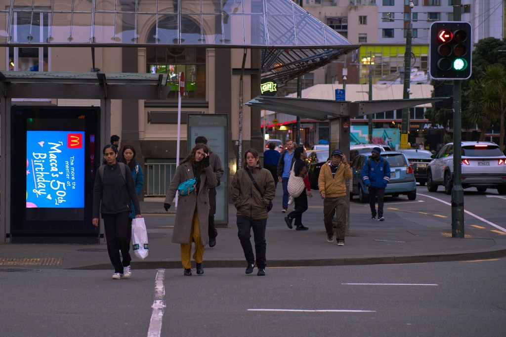 A busy urban street scene with pedestrians crossing an intersection near a bus stop, featuring a colorful McDonald's advertisement celebrating its 50th birthday.