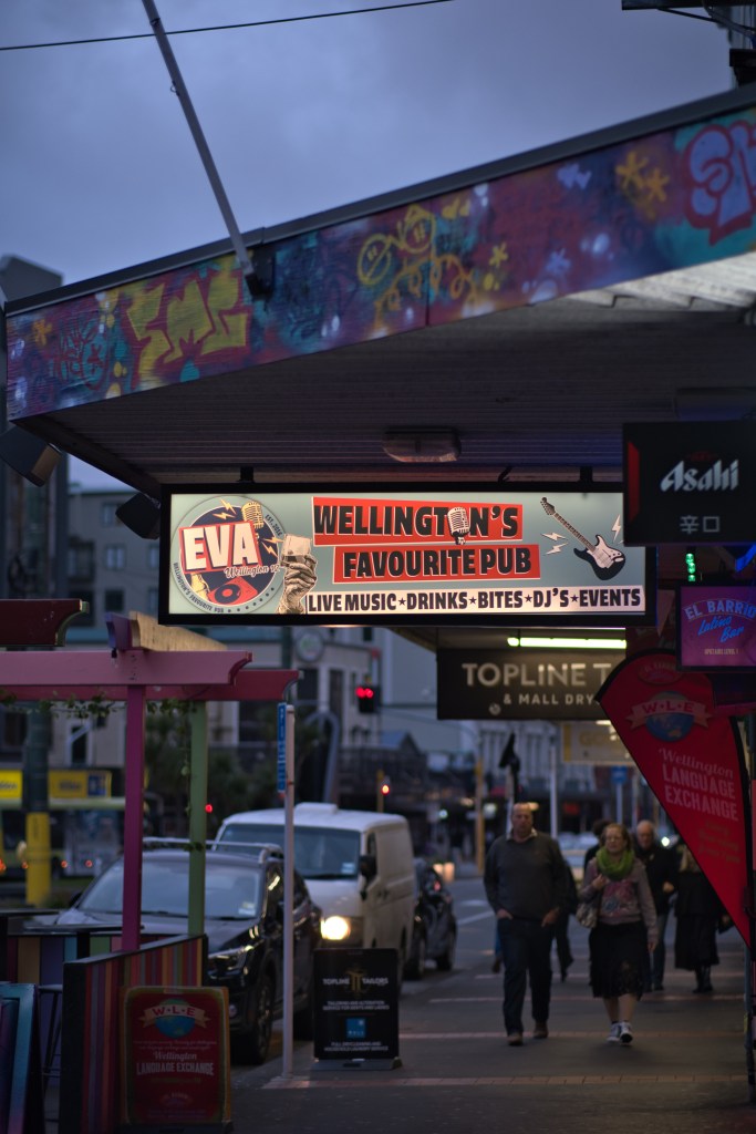 A brightly lit sign for 'Eva', a pub in Wellington, featuring 'Wellington's Favourite Pub' text, promoting live music, drinks, bites, DJs, and events, with pedestrians walking by and colorful storefronts in the background.