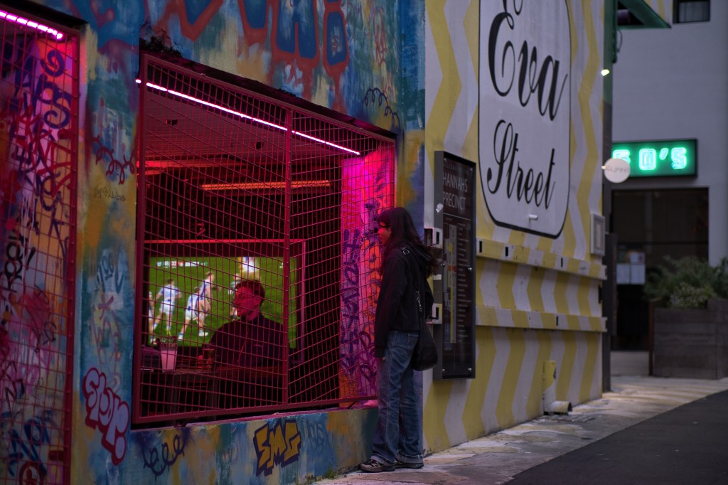 A person stands in front of a colorful graffiti-covered wall with pink neon lights, looking into a wired enclosure where another individual is seated, watching a screen. The scene captures an urban environment with street art and an illuminated sign reading 'Eva Street.'
