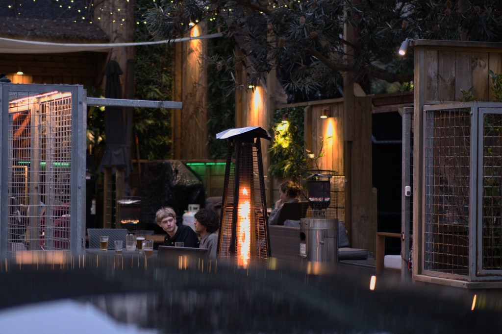 A cozy outdoor seating area in the evening, featuring two people conversing near a heater, surrounded by wooden structures and soft lighting.