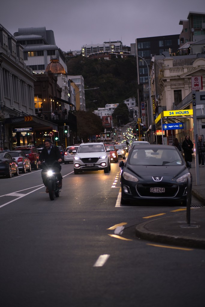 A busy city street in the evening, featuring cars and a motorcycle, with buildings lining the sides and a hill in the background under a cloudy sky.