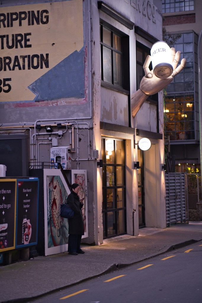 A woman standing outside a bar with a large hand sculpture holding a beer can above the entrance. The building features graffiti and a colorful mural on the wall.