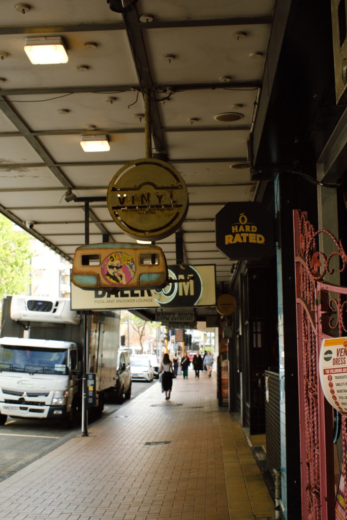 A street view featuring various business signs including 'VINYL' and 'BILLIARD ROOM', with pedestrians walking along a tiled sidewalk.
