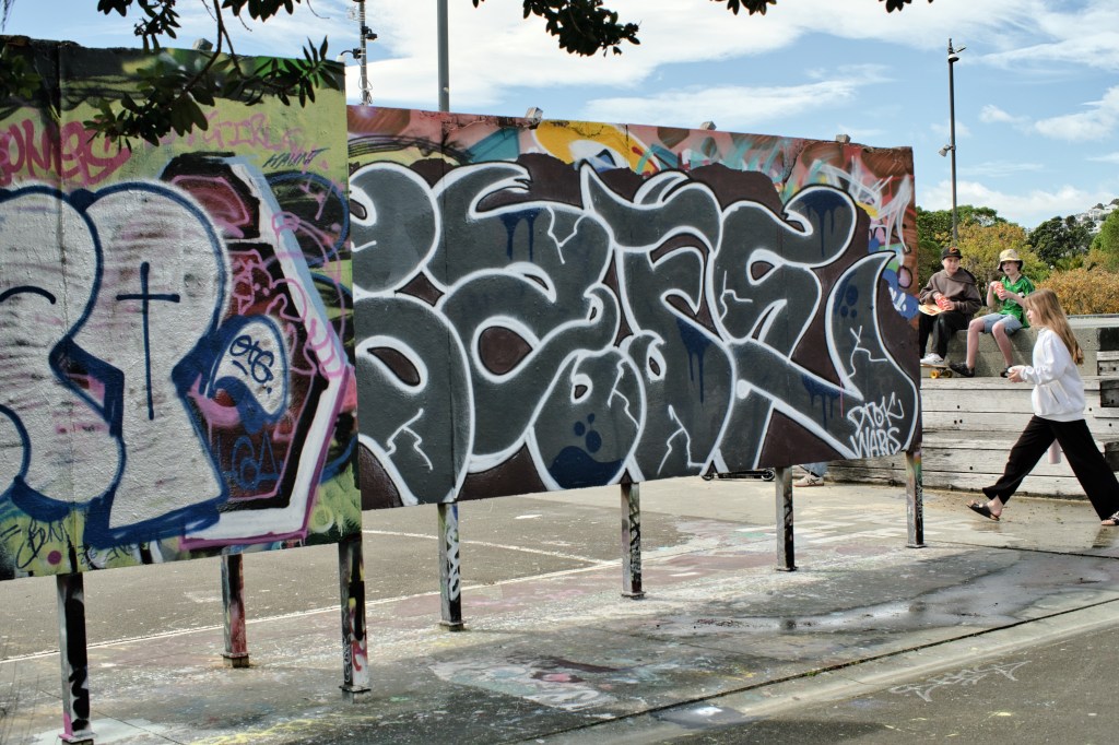 Colorful graffiti on large boards with people sitting on a ledge in the background under a blue sky.