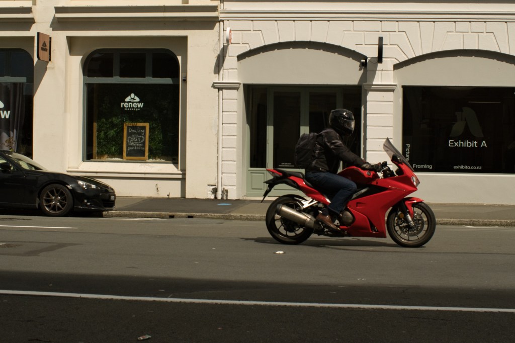 A motorcyclist riding a red motorcycle on a city street, in front of shops and parked cars, under clear skies.