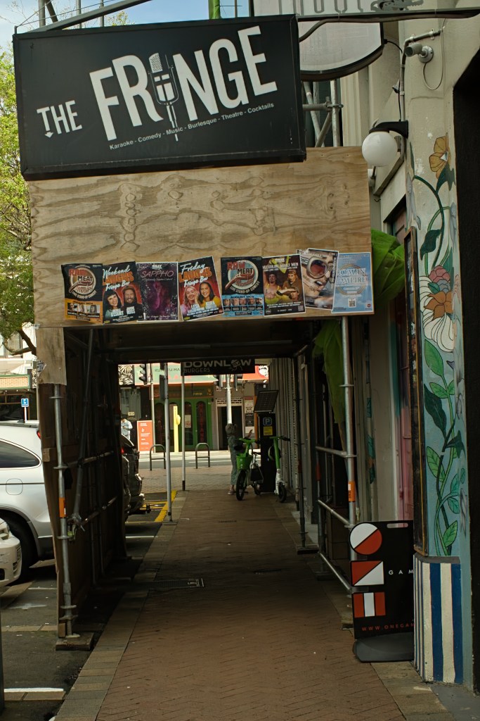 Entrance to The Fringe venue showcasing a sign with various events and entertainment options, surrounded by scaffolding and murals on the walls.