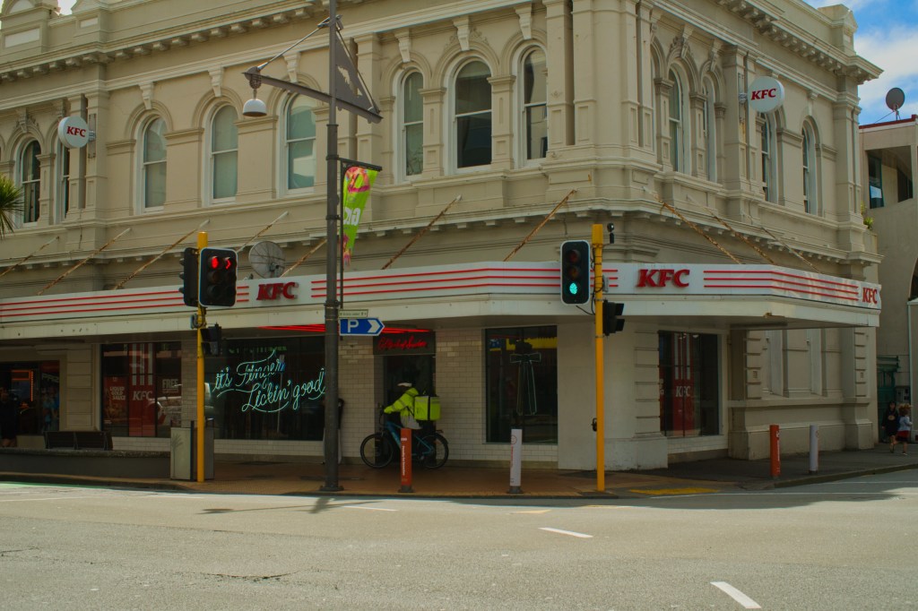 Exterior view of a KFC restaurant located in a historic building, featuring its iconic signage and a vibrant street scene with traffic signals and pedestrians.