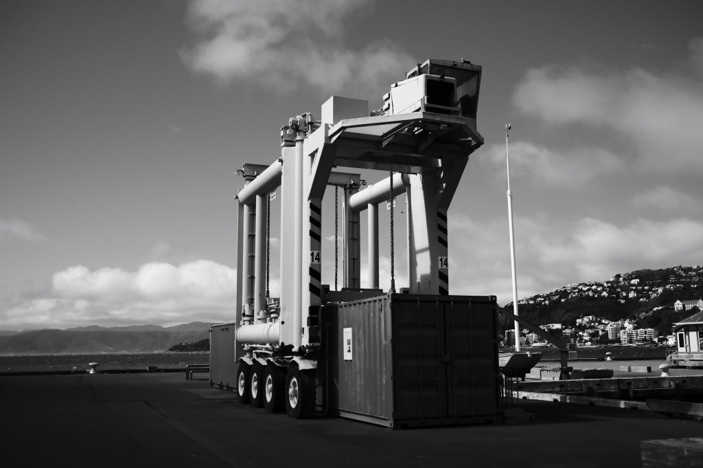 A large container crane positioned at a port, with several cylindrical structures and wheels, set against a backdrop of clouds and distant hills.