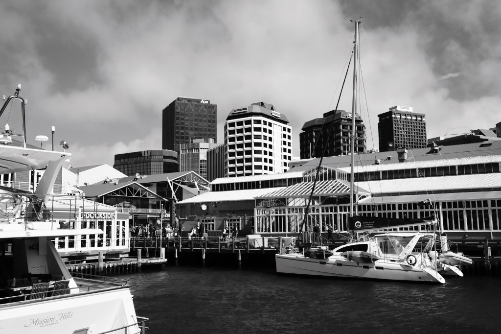 Black and white image of a harbor with boats docked, surrounded by modern skyscrapers and a cloudy sky.