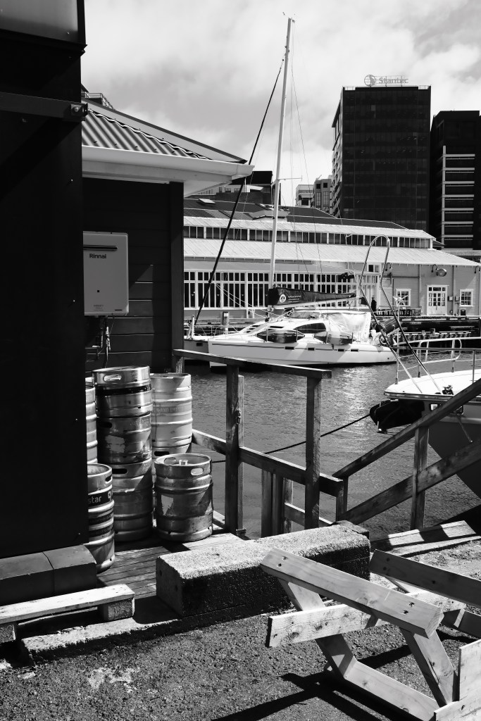 Black and white image of a marina with moored sailboats and a building featuring stacked kegs in the foreground.
