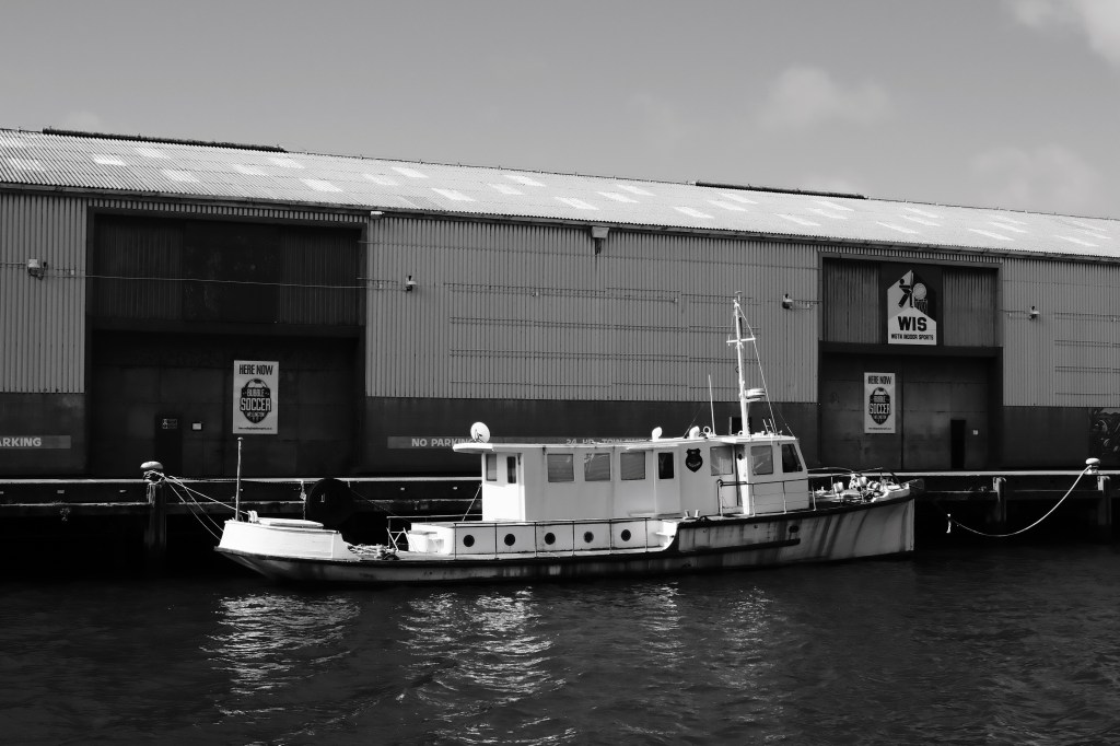 A white boat docked at a pier in front of a large warehouse, captured in black and white. The warehouse features signage promoting soccer and sports activities.