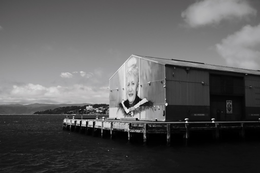 A black and white photo of a waterfront warehouse featuring a large mural of a smiling woman. The structure is located on a pier with a view of the sea and distant hills.