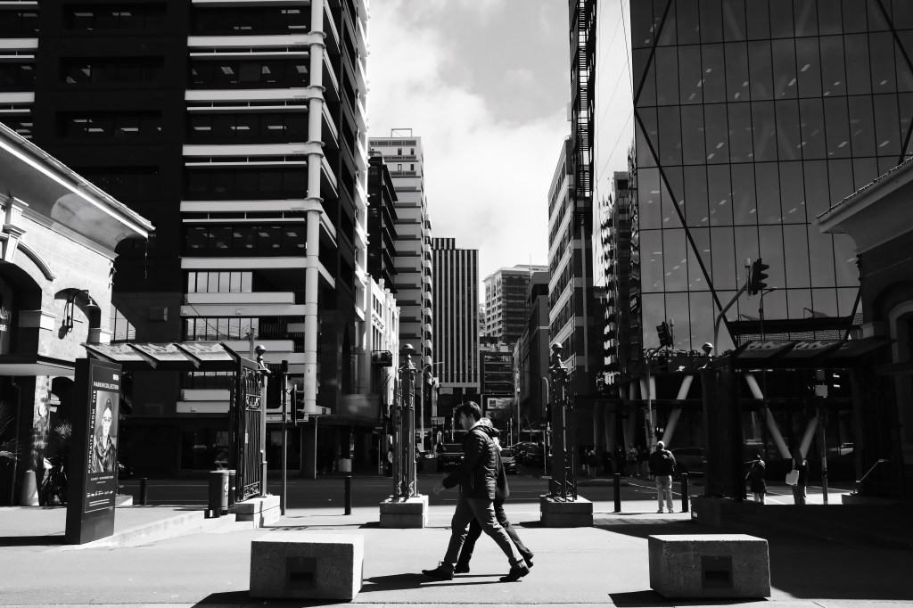 A person walking through a city street lined with tall buildings, featuring a mix of modern glass structures and older architecture, captured in black and white.