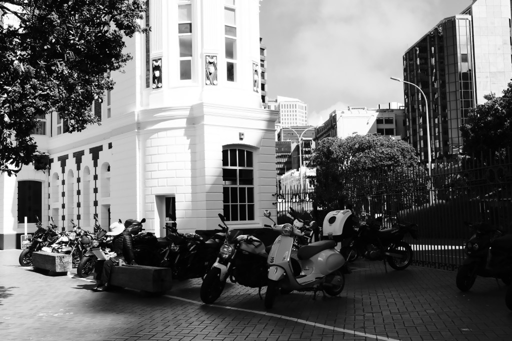 Black and white image of a street scene with a parked row of motorcycles beside a historic building, featuring a person sitting on a bench nearby.