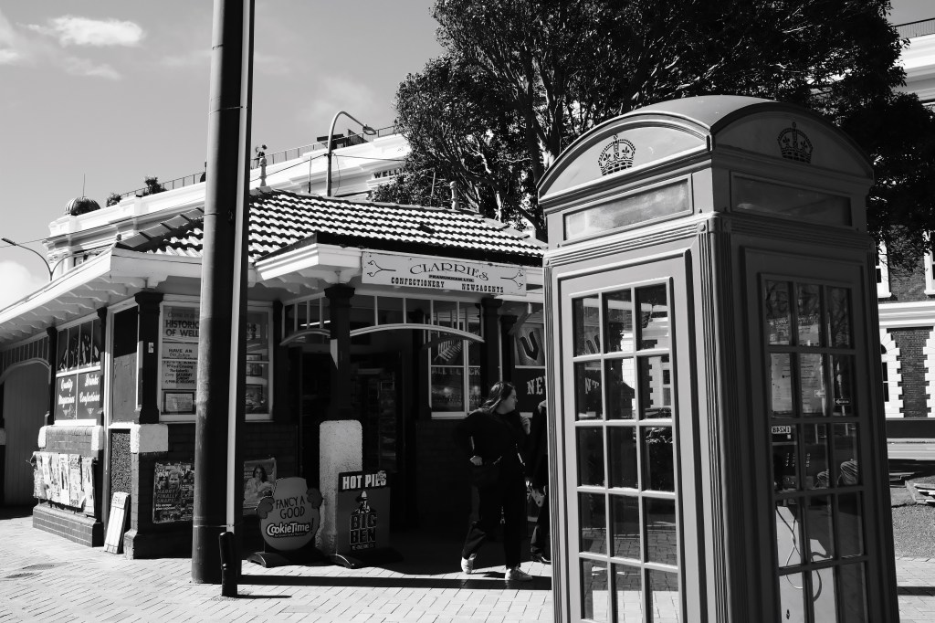A black and white image of a historic building with a striped roof, featuring a sign that says 'Clardee's Confectionery'. In the foreground, there is a traditional red telephone booth, with a woman standing nearby.