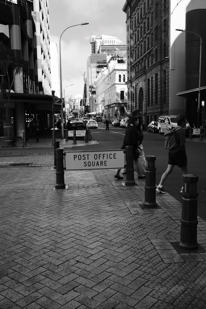 Black and white street view with a sign for 'Post Office Square' and pedestrians crossing a road lined with buildings.